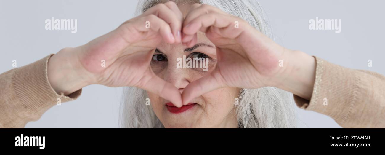 Flirtende alte Frau sieht durch ein Herz aus Fingern Stockfoto