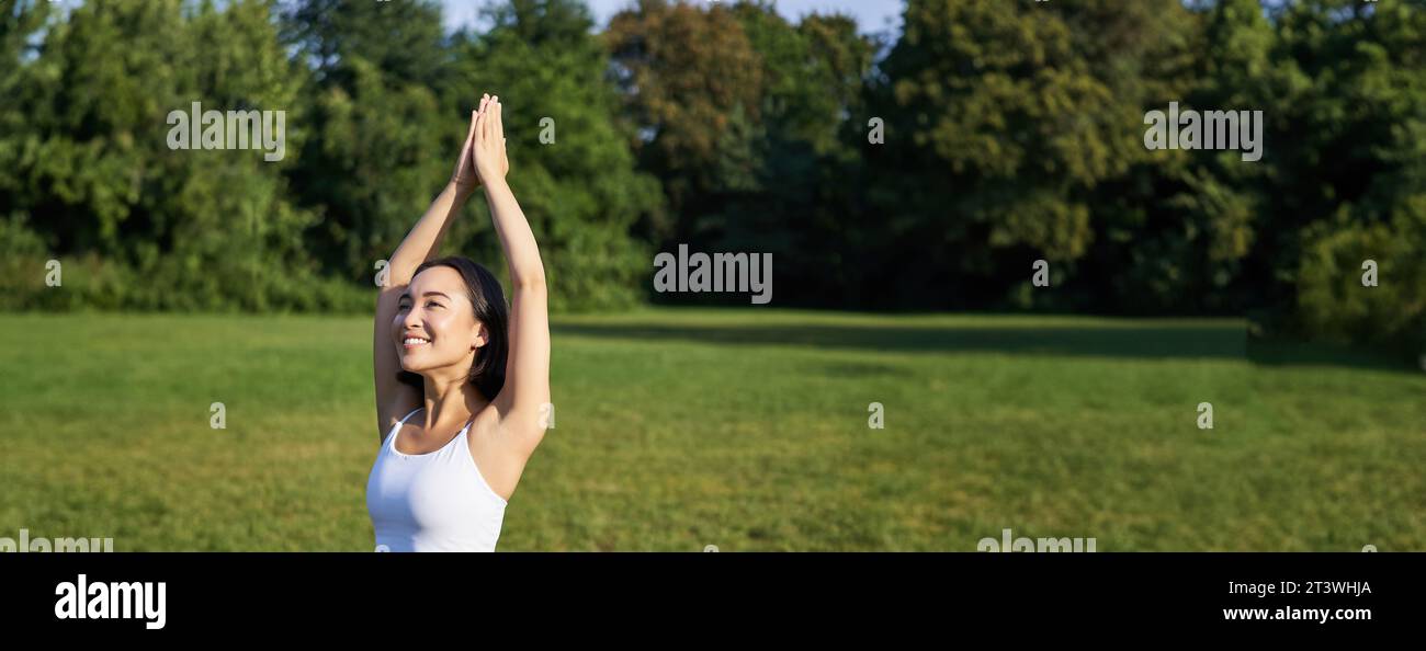 Vertikale Aufnahme eines jungen asiatischen Fitness-Mädchens, das Yoga Asana macht, in Baumhaltung steht und lächelt und im Park auf einer Gummimatte ausübt Stockfoto