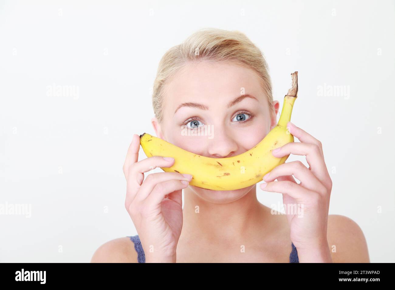 Junge weibliche blonde kaukasische Modell vor weißem Hintergrund, die gesunde Banane als Teil eines Smiley Face hält - gesunde Ernährung Konzept Stockfoto