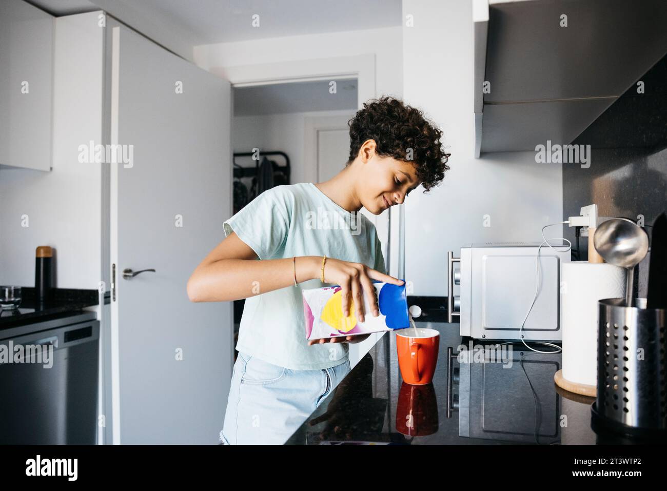 Zehn Jahre altes Mädchen, das Milch in einen roten Becher in der Küche gießt. Ein Mädchen zu Hause, das eine Tasse Milch zubereitet. Stockfoto