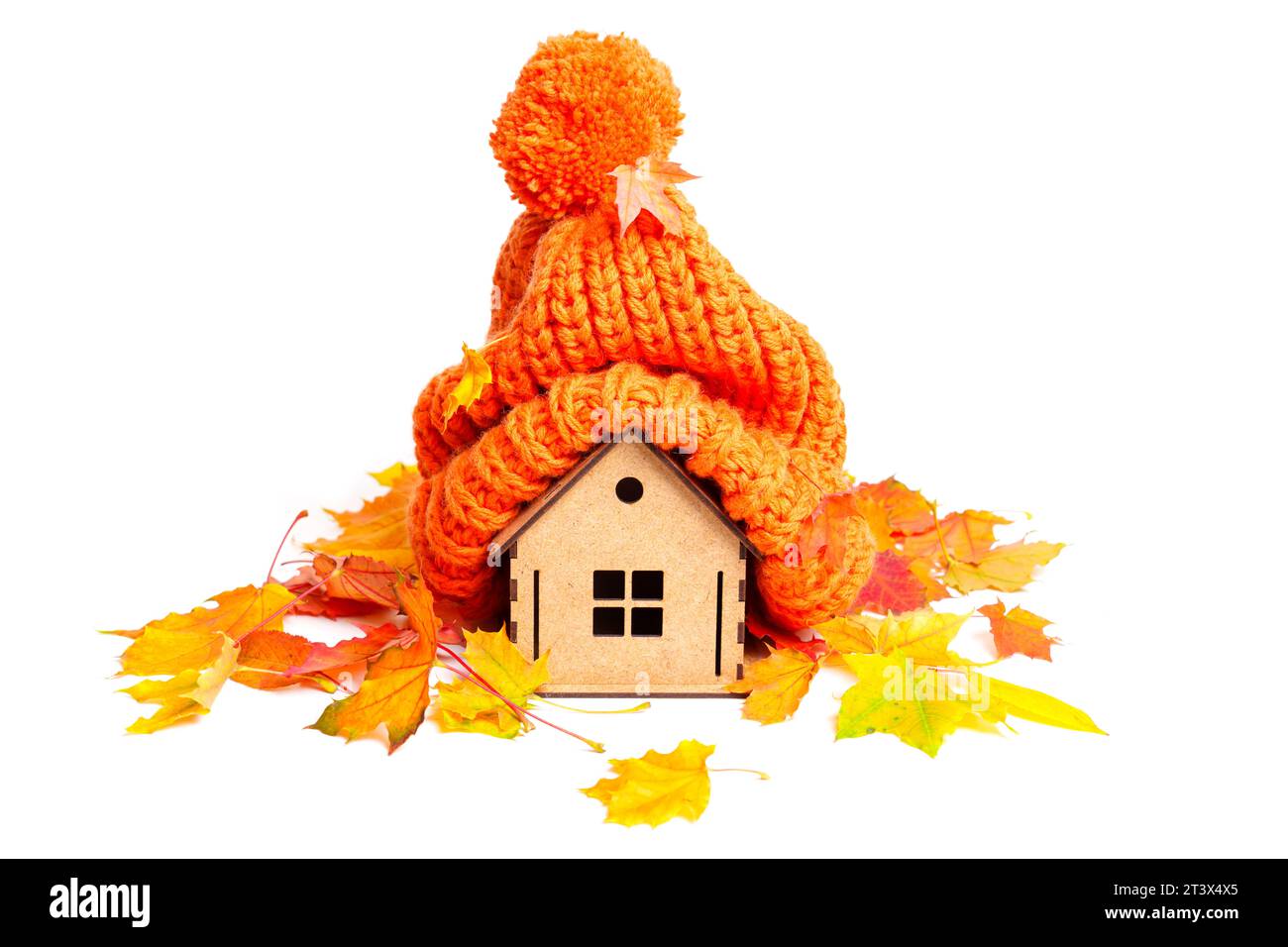 Bezauberndes Miniatur-Holzhaus mit einem warmen, orangen Strickmütze mit Herbstlaub auf weiß. Tragekomfort und Wärme in der kalten Jahreszeit Stockfoto