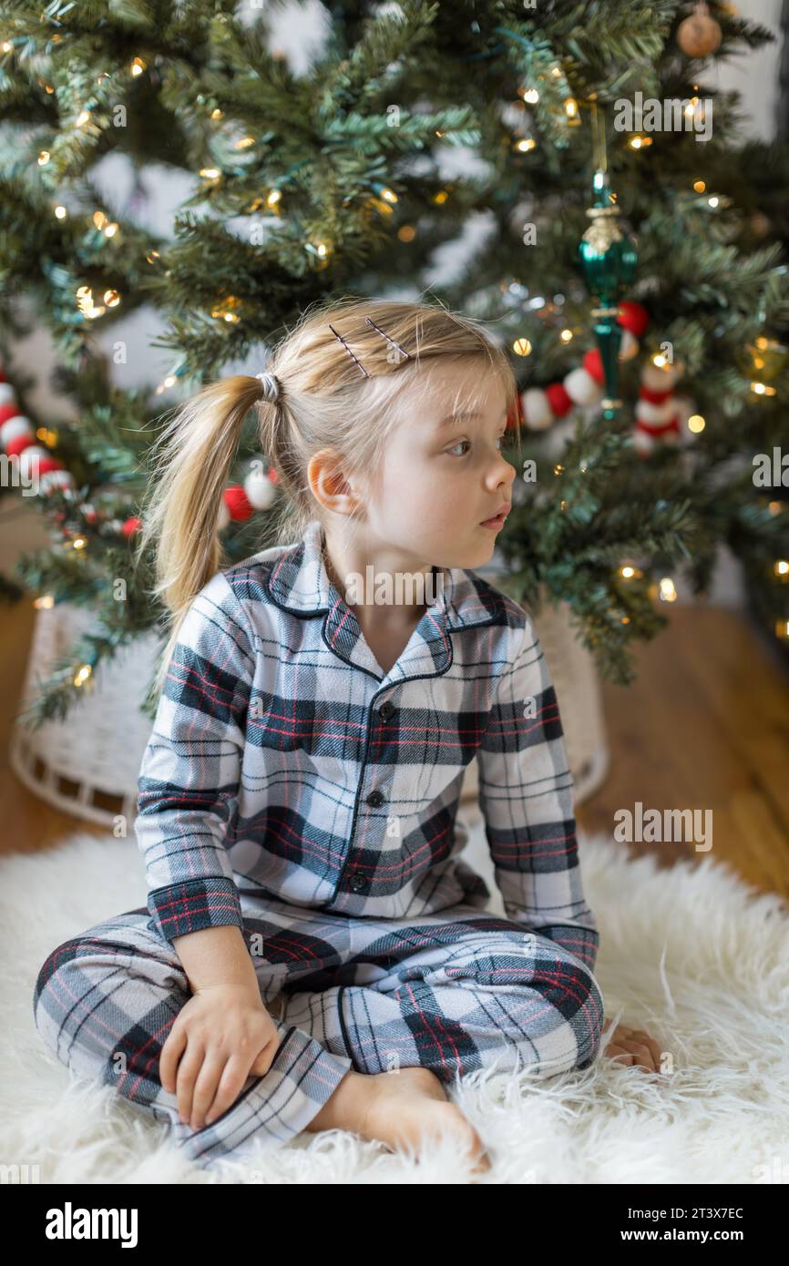 Neugieriges Kleinkind im karierten Pyjama sitzt am Weihnachtsbaum Stockfoto