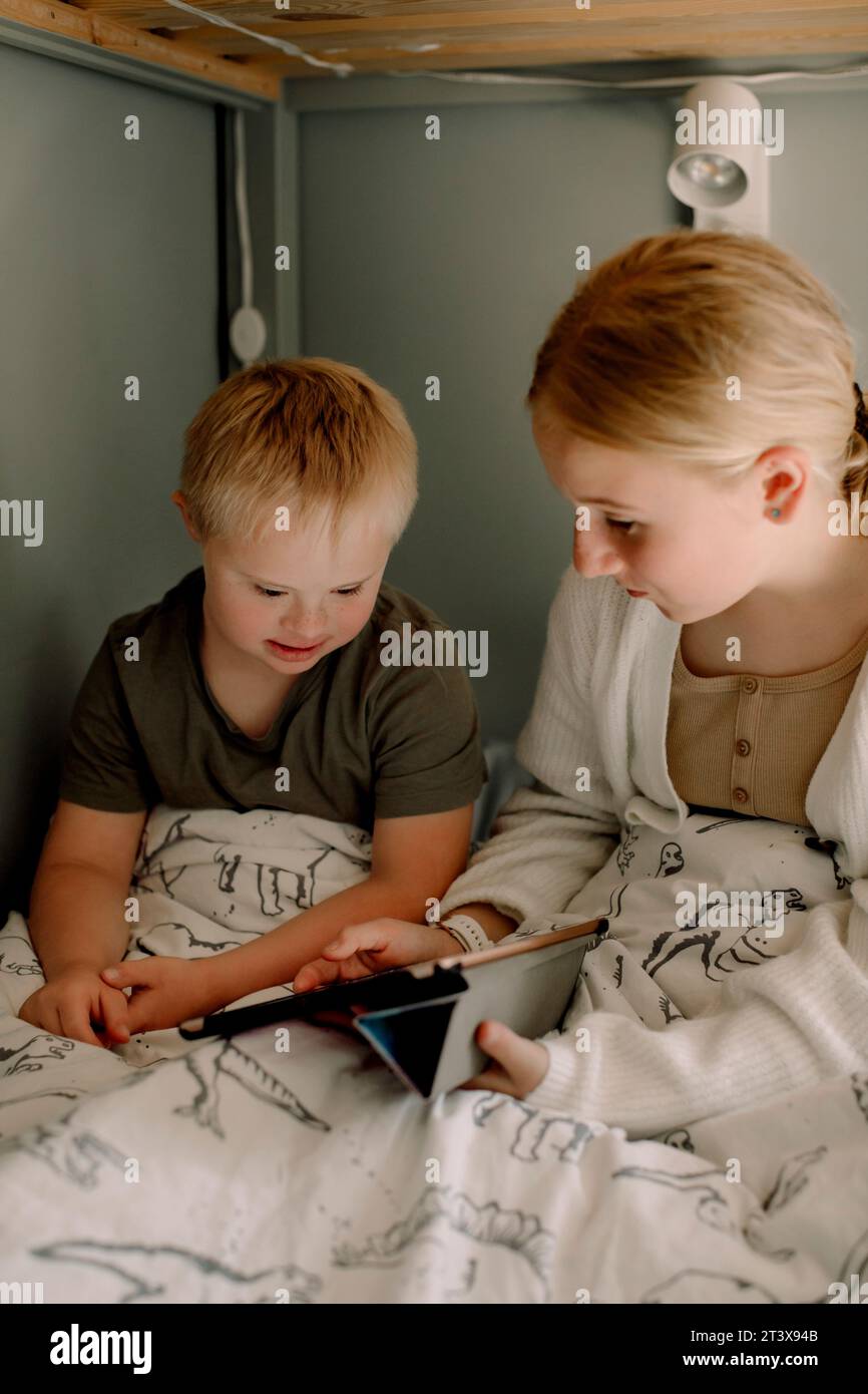 Ein Mädchen teilt sich ein digitales Tablet mit einem Bruder, der zu Hause auf dem Bett sitzt Stockfoto