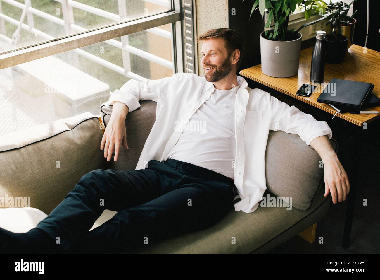 Blick aus einem hohen Winkel auf einen lächelnden, durchdachten Geschäftsmann, der sich im Büro auf dem Sofa zurücklehnt Stockfoto