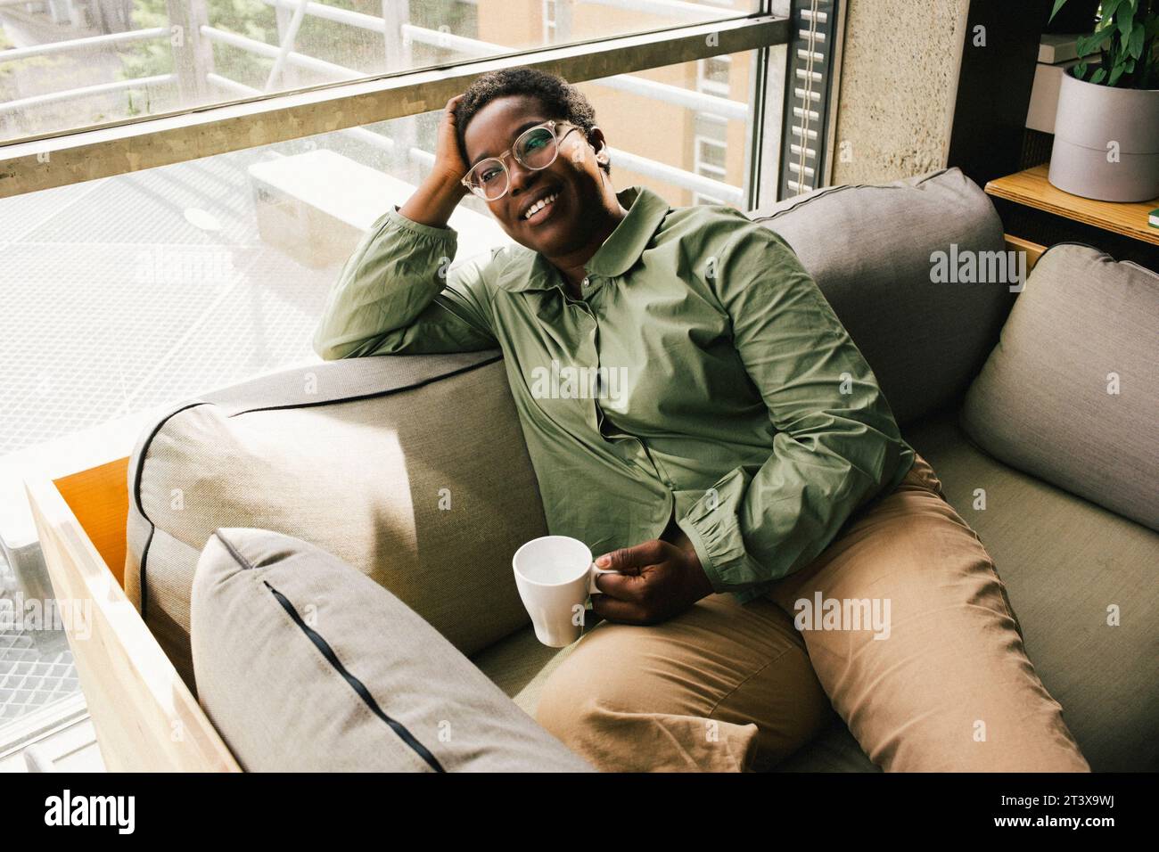 Lächelnde Geschäftsfrau, die mit einer Kaffeetasse sitzt, während sie tagsüber im Büro träumt Stockfoto
