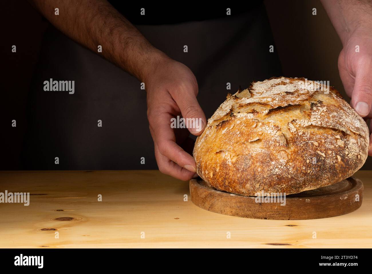 Die Hände eines jungen Mannes, der Sauerteigbrot behandelt und das Brot mit schönen goldenen Tönen vor dem dunklen Hintergrund hervorhebt. Stockfoto