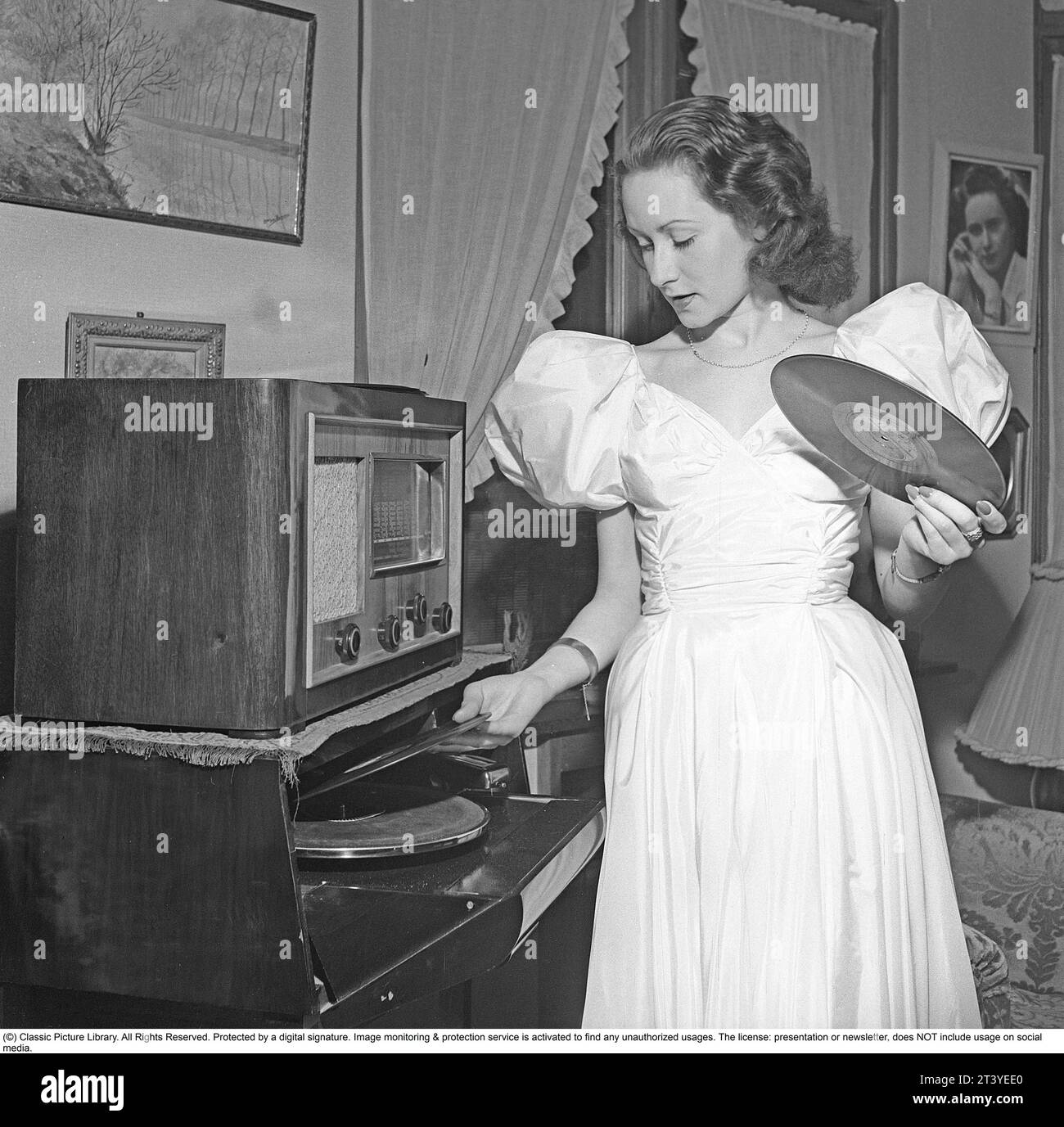 Frau am Gramophon in den 1940ern Sie legt noch eine Platte auf den Teller, um sie zu hören. Die Platten waren aus zerbrechlichem Material und die Geschwindigkeit der Aufnahme, wie sie sollte, betrug 78 Umdrehungen pro Minute. Ein Funkgerät steht auf dem Grammophon. Schweden 1947. Kristoffersson Ref. V149-3 Stockfoto