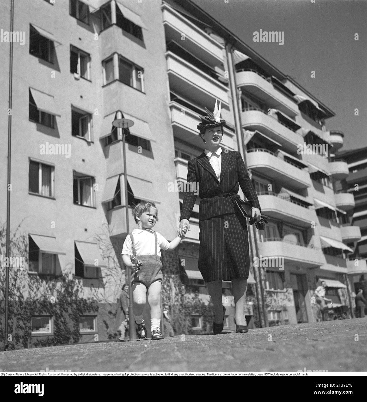 Einen Tag im Mai 1940. Eine Mutter hält ihren Sohn in der Hand, während sie auf einer Straße in Stockholm spaziert. Im Hintergrund ein Wohnhaus und Wohnungen. Sie ist in typischer 40er-Mode gekleidet. Ein Kleid mit Kreidestreifen, passendem Rock, Jacke und wunderschöner Hut. Schweden Stockholm Mai 1940. Kristoffersson Ref. 133-21 Stockfoto