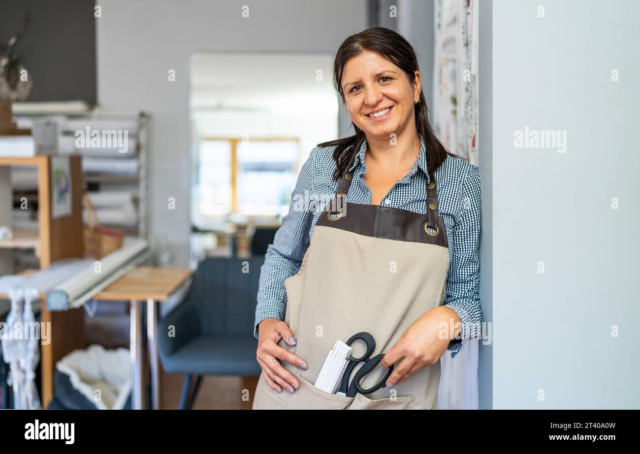 Eine lächelnde Schneiderin in einem karierten Hemd und einer beigefarbenen Schürze steht an einem Arbeitsplatz und hält eine Schere und ein Maßstabslineal in der Schürzentasche Stockfoto