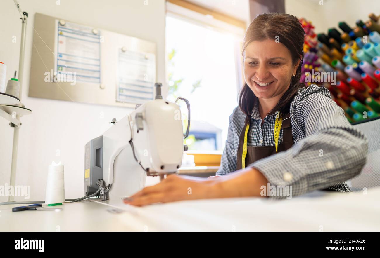 Fröhliche Frau in einer Schürze arbeitet an einer Nähmaschine und näht weißen Stoff in einem gut beleuchteten Arbeitsbereich mit bunten Fadenspulen im Hintergrund Stockfoto