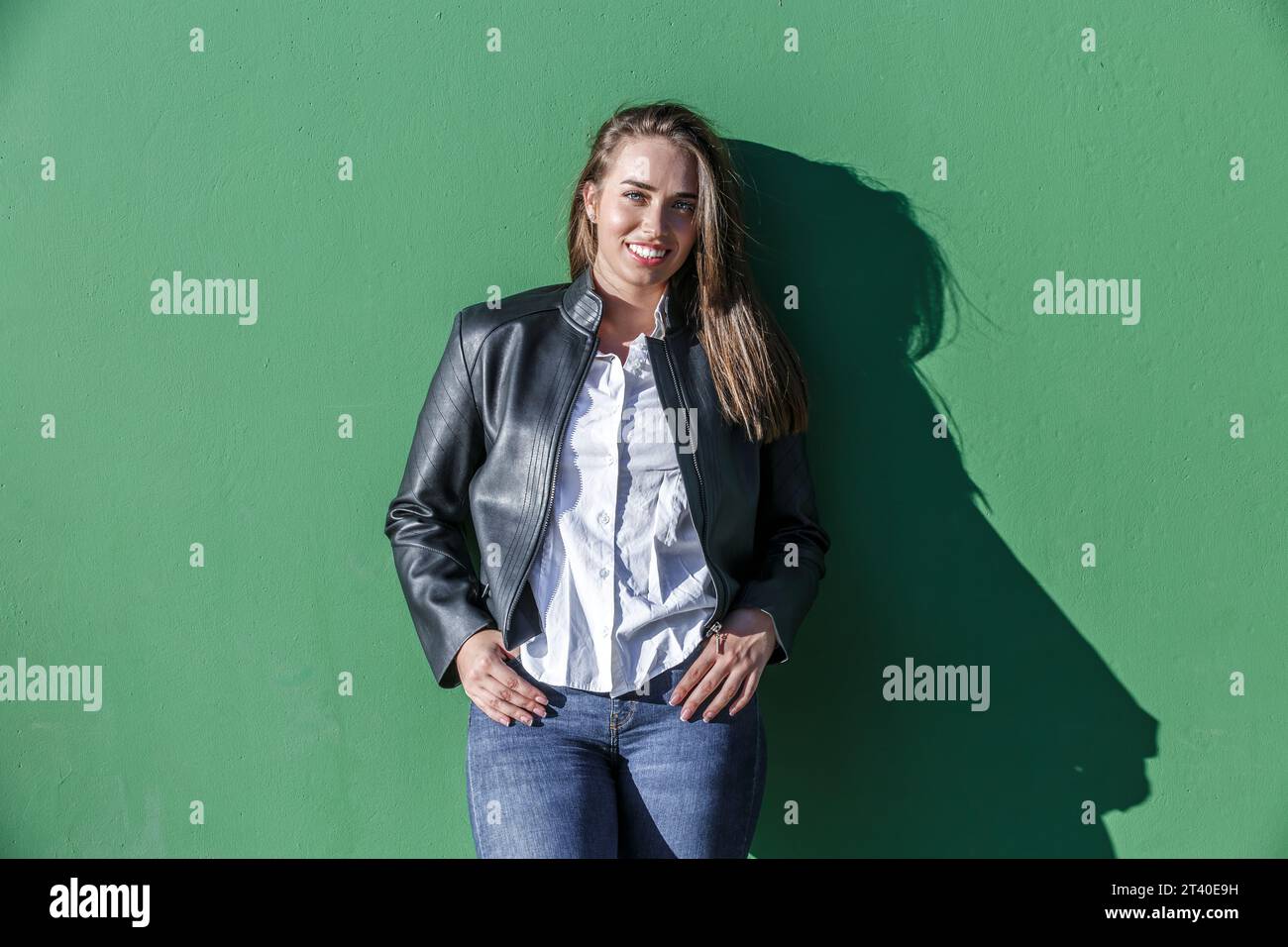 Selbstbewusste junge, lächelnde Frau mit Lederjacke und Jeans, die Finger in die Taschen steckt, während sie vor der grünen Wand mit Schatten des Sonnenlichts steht Stockfoto