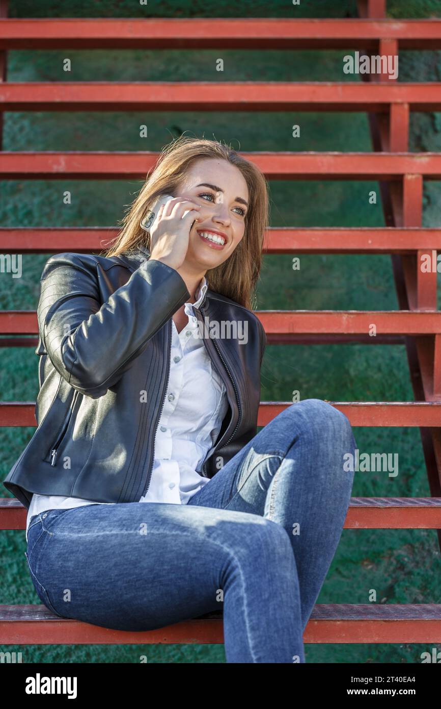 Junge lächelnde Frau in legeren Kleidern, die auf einer roten Treppe sitzt und mit einem Freund während eines Telefonanrufs auf der Straße im Sonnenlicht spricht Stockfoto