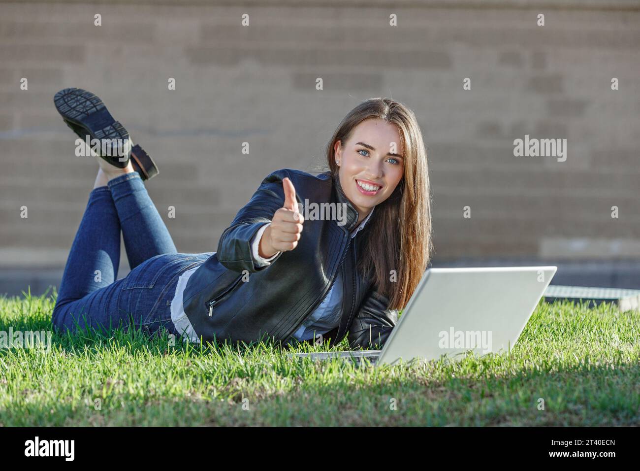 Junge glückliche Studentin in lässigem Outfit, die auf dem Bauch mit den Füßen nach oben auf dem Rasen liegt und Netbook durchsucht, während sie wie Gesten zeigt und in die Kamera schaut Stockfoto