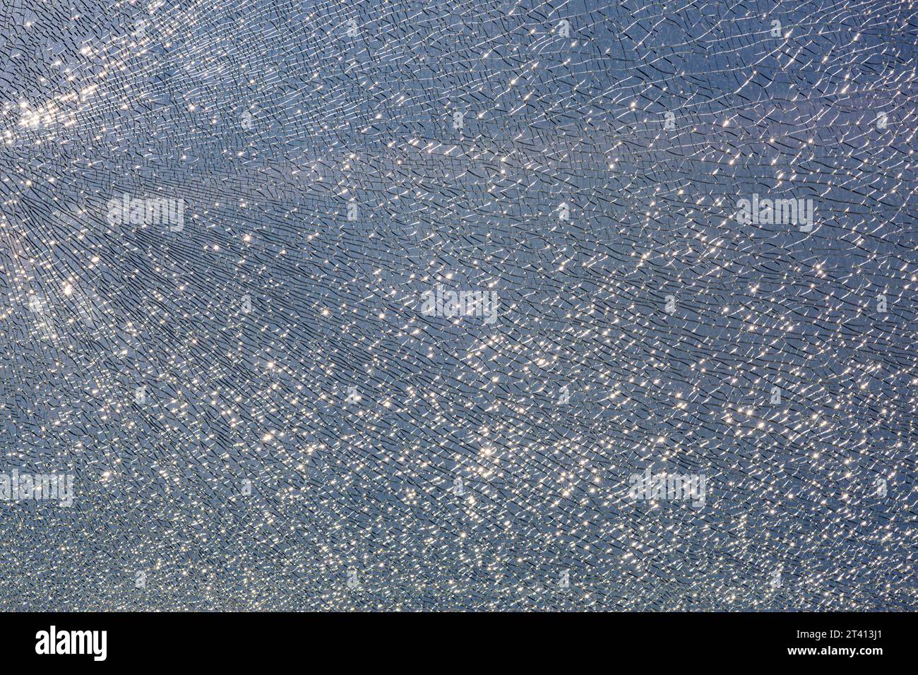 Gebrochenes gehärtetes Glas in Rissen, verdorbene Glasoberflächentextur auf blauem Himmel Hintergrund, Glanz von hellem Licht, Nahaufnahme. Stockfoto