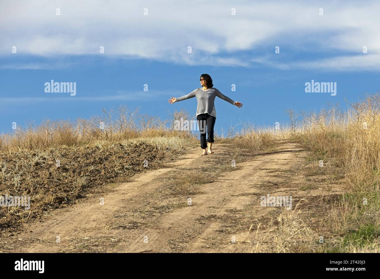Eine asiatische Frau mit Sonnenbrille geht barfuß auf einem Feldweg neben einem Feld in Idaho und bekommt die Vorteile der Erdung. Stockfoto