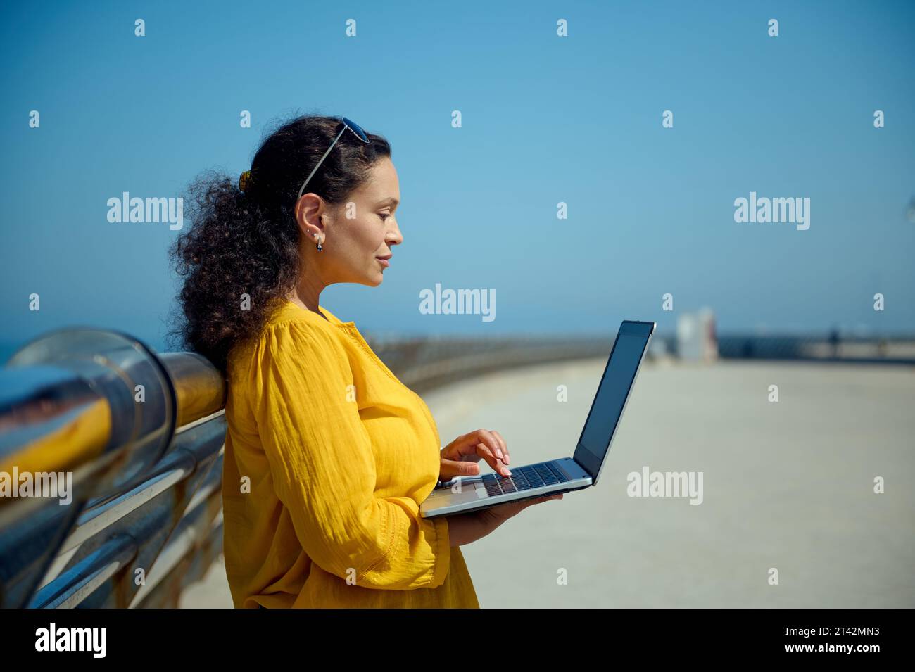 Seitenporträt einer Multi-ethnischen schönen Frau mittleren Alters, Geschäftsfrau, freiberuflicher Unternehmer in gelbem Hemd, mit Laptop, Working remo Stockfoto