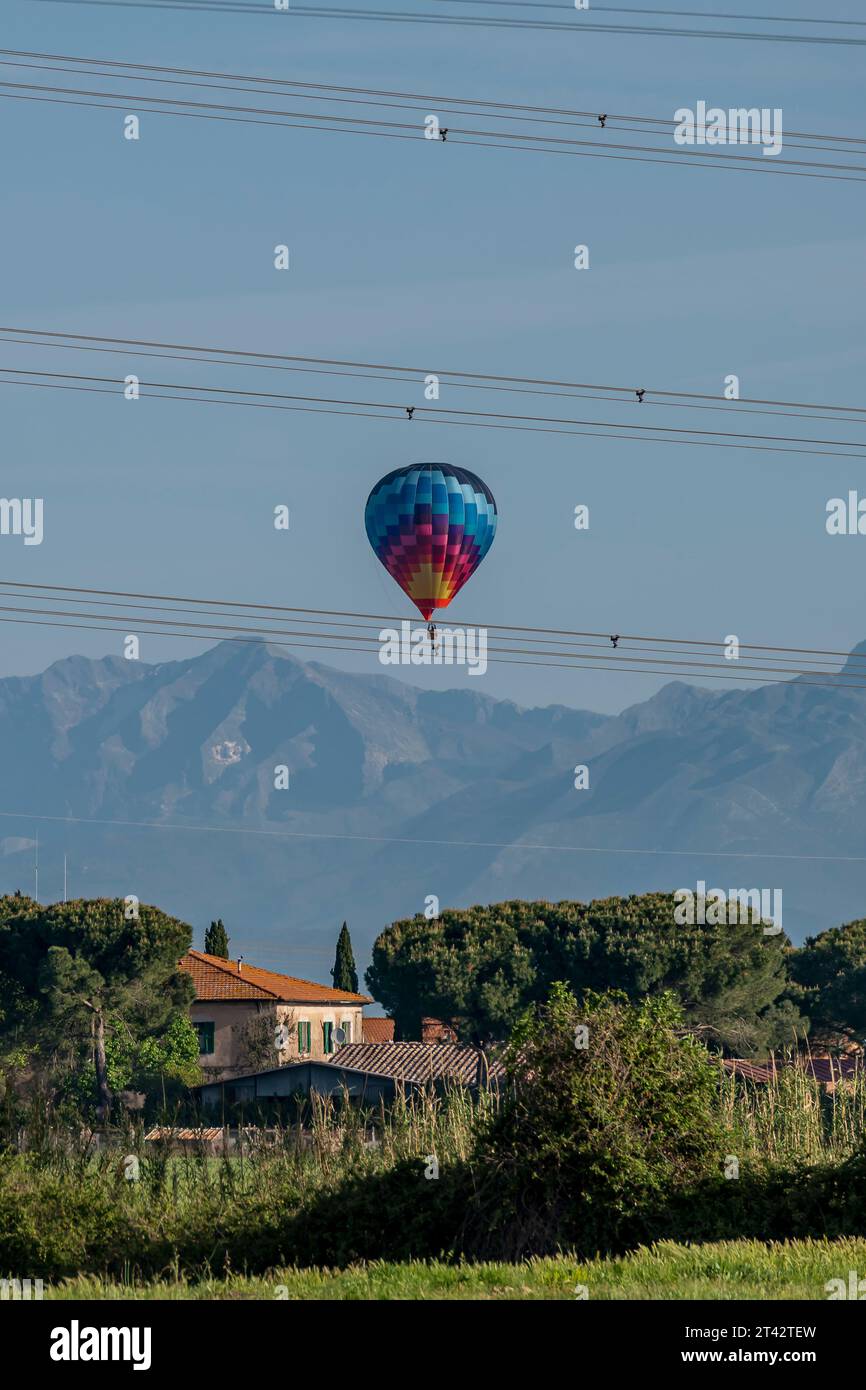 Ein bunter Heißluftballon fliegt über ein Haus und Hochspannungsdrähte, mit Bergen im Hintergrund, Fauglia, Italien Stockfoto