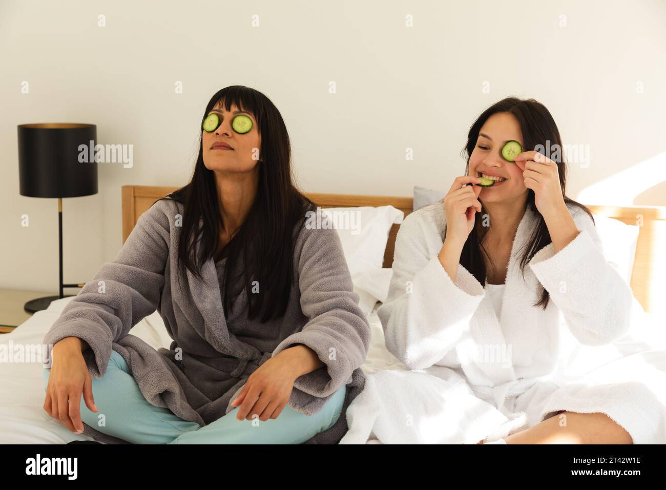 Diverse Mutter und Tochter in Bademänteln, sitzen auf Bett mit Gurken auf Gesichtern zu Hause, Kopierraum Stockfoto