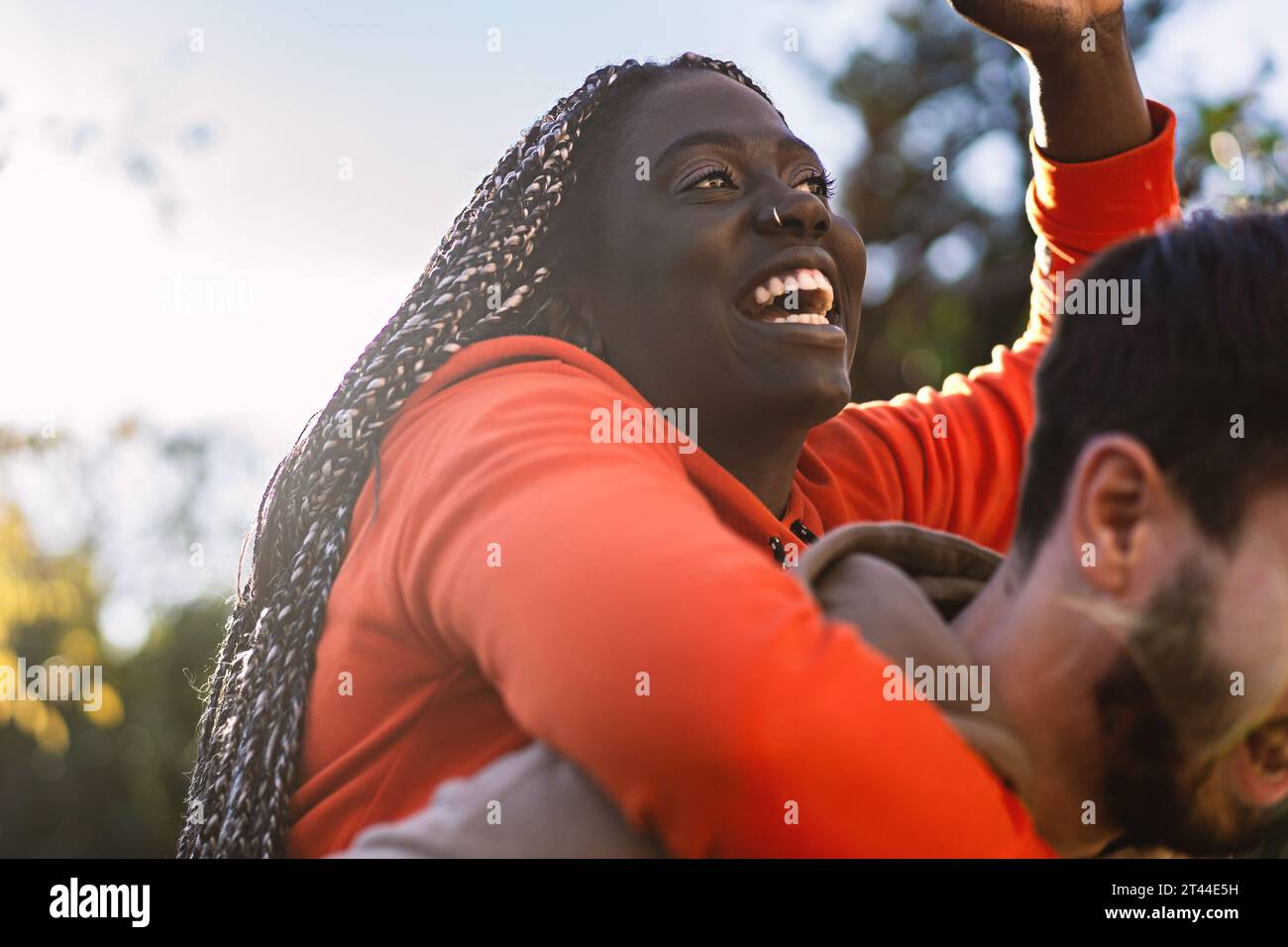 Die fröhliche afrikanische Abstammungsfrau mit langen Zöpfen lacht fröhlich, während sie von ihrem Partner im Freien einen Huckepack-Ritt bekommt, um einen echten festzuhalten Stockfoto