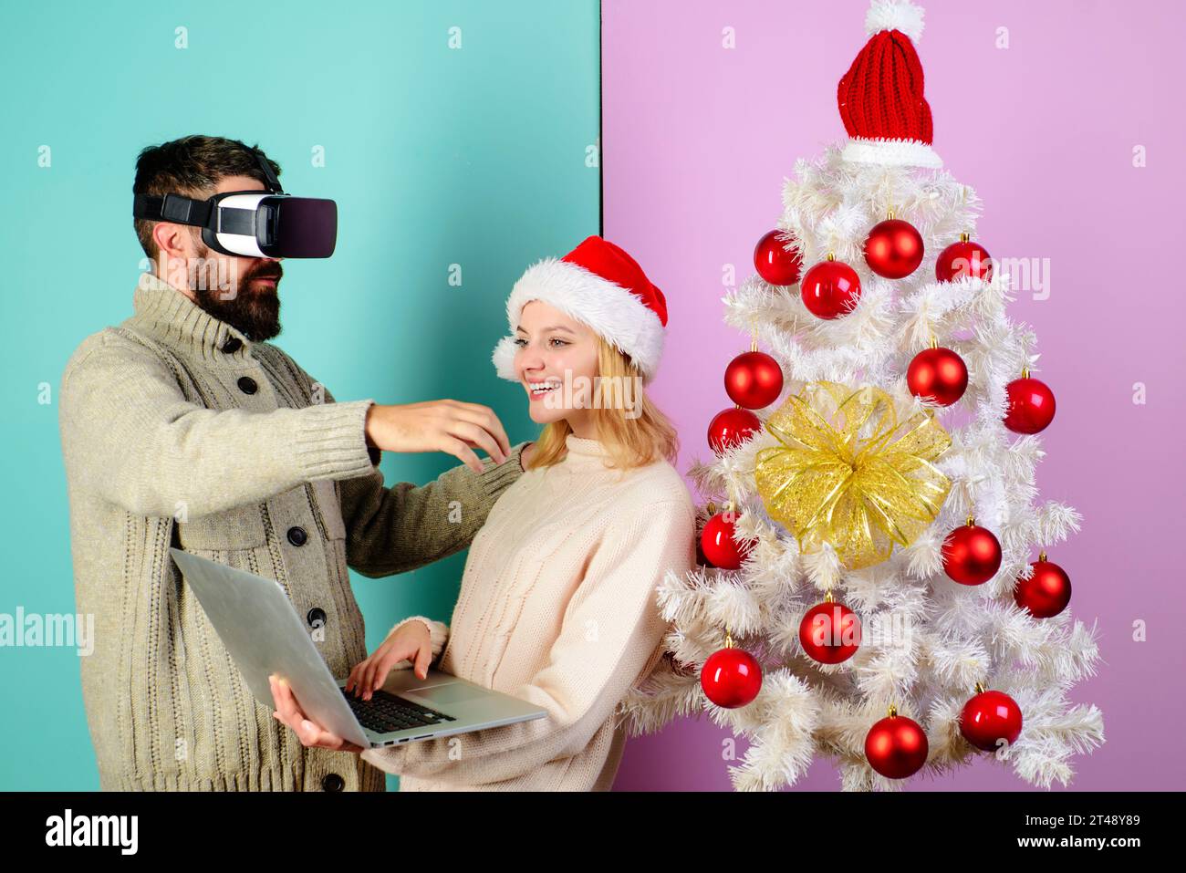 Glückliche Familie in der Nähe des Weihnachtsbaums. Bärtiger Mann in VR-Brille und lächelnde Frau in Santa-Hut mit Laptop. 3D-Technologie, Unterhaltung. Weihnachtspaar Stockfoto