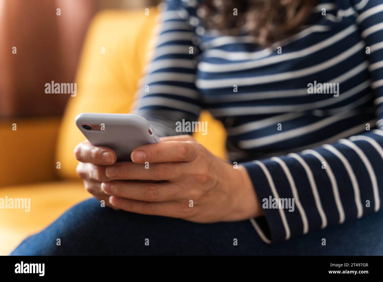 Ein Mädchen in Jeans und einer gestreiften Bluse sitzt auf dem Sofa und blättert durch die Seiten auf ihrem Smartphone-Bildschirm, um unseren Wunsch zu verkörpern, immer auf dem neuesten Stand zu sein Stockfoto