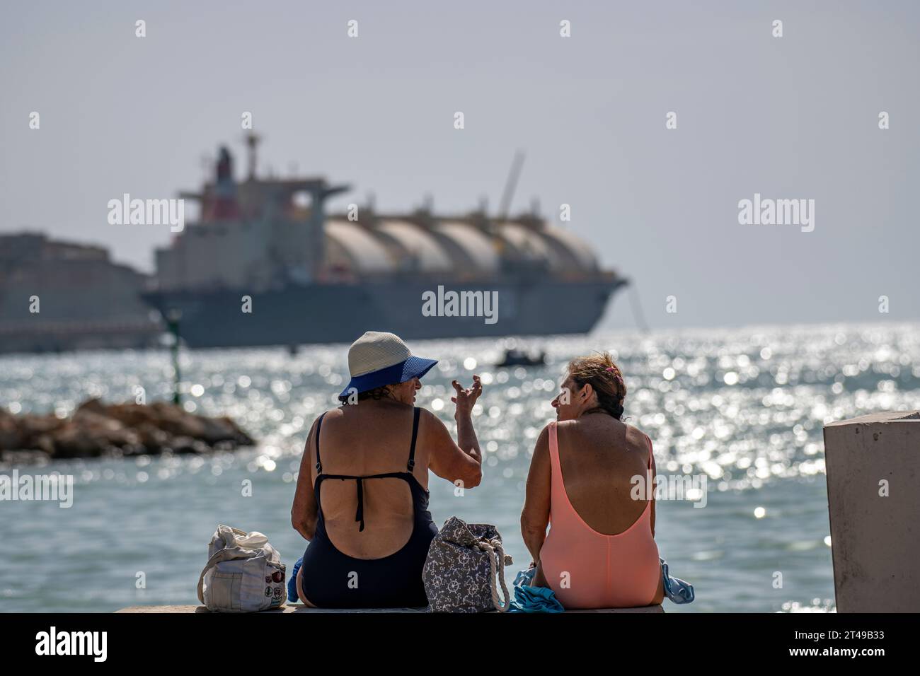 Zwei Damen in Badeanzügen genießen den Strand mit der Armada LNG Mediterrana in der Ferne, einer schwimmenden Gasspeicher- und -Rückvergasungseinheit in Malta. Stockfoto