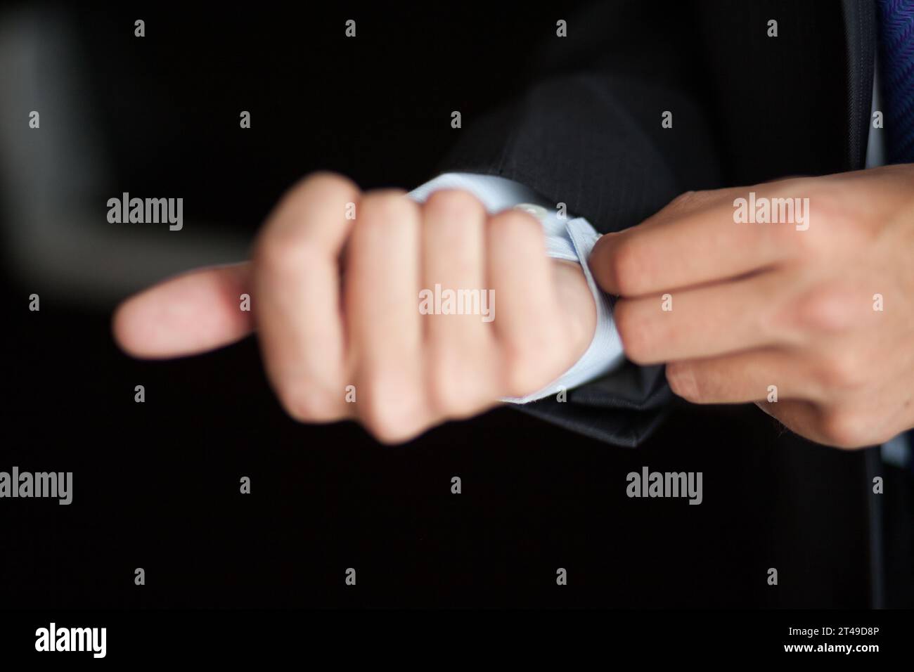 Nahaufnahme eines Mannes, der ein Hemdbündchen anpasst, mit einem kontrastierenden dunklen Anzug. Fokussiert auf die Hände, mit verschwommenem Hintergrund. Stockfoto