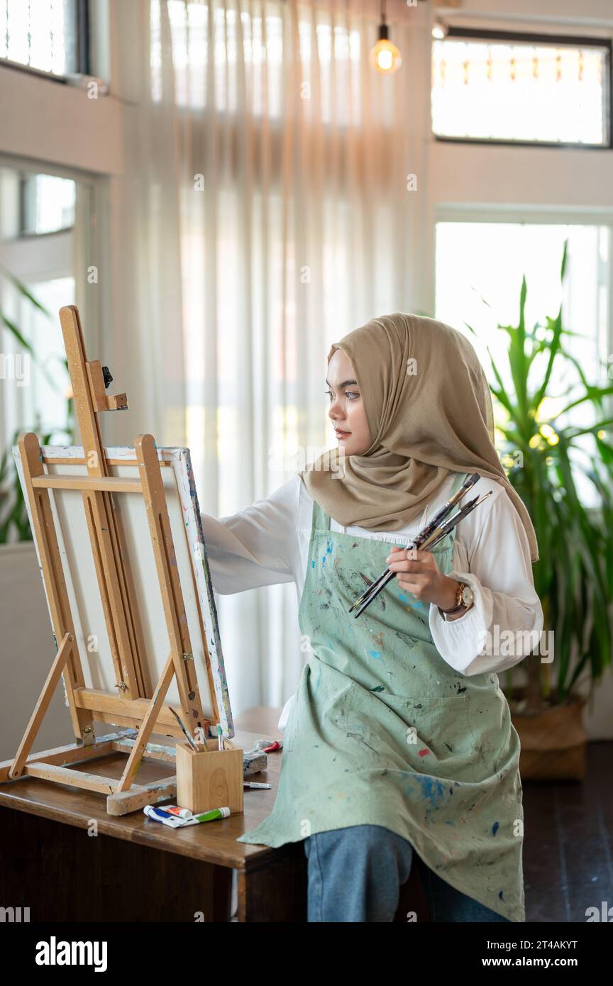 Eine kreative und fokussierte asiatisch-muslimische Künstlerin auf einer Schürze malt auf Leinwand auf einer Staffelei und arbeitet in ihrem Atelier. Stockfoto