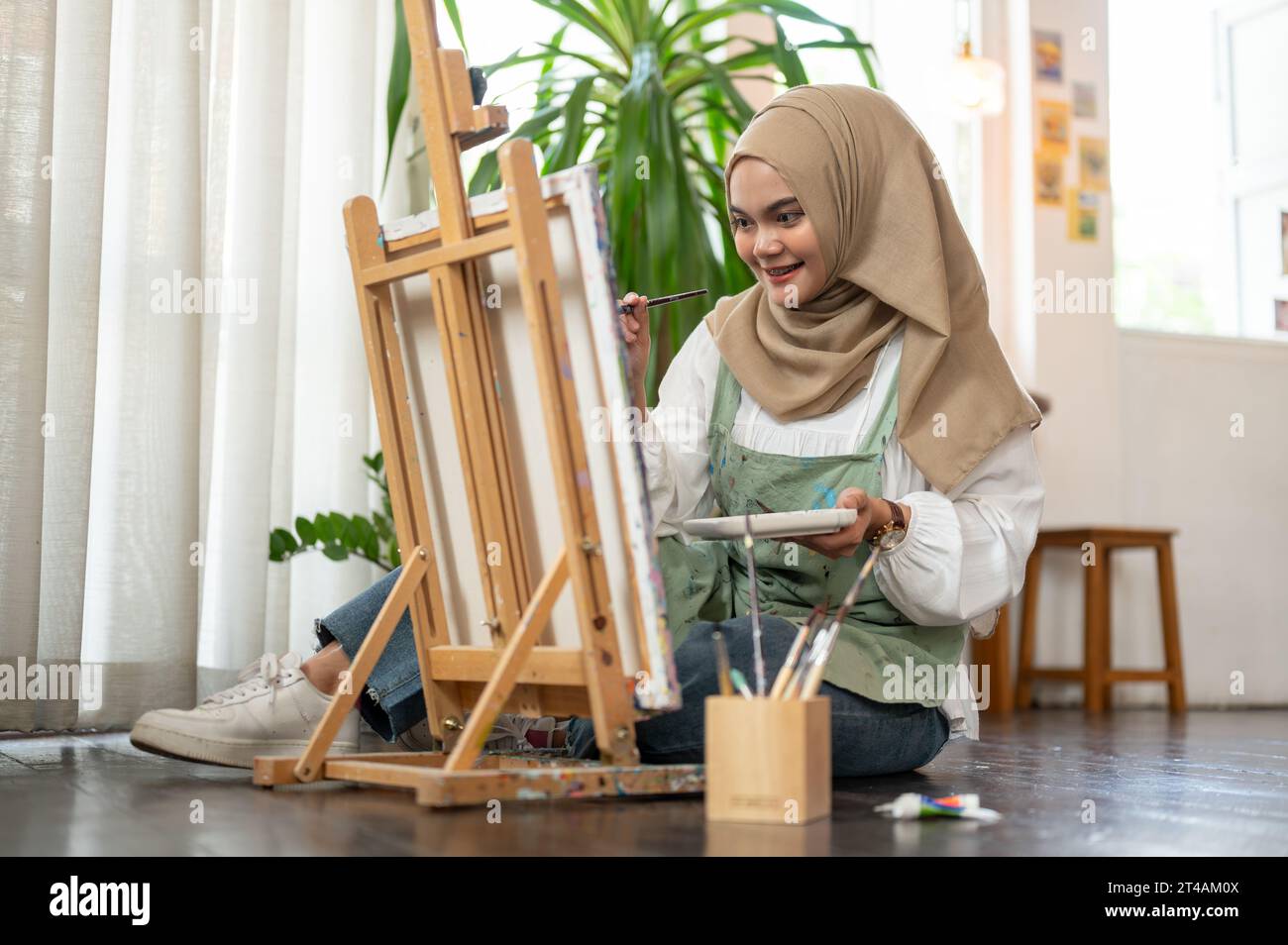 Eine fröhliche und kreative asiatisch-muslimische Künstlerin in einer Schürze sitzt in ihrem Kunststudio auf dem Boden, zeichnet oder malt auf Leinwand auf einer Staffelei und arbeitet in Stockfoto