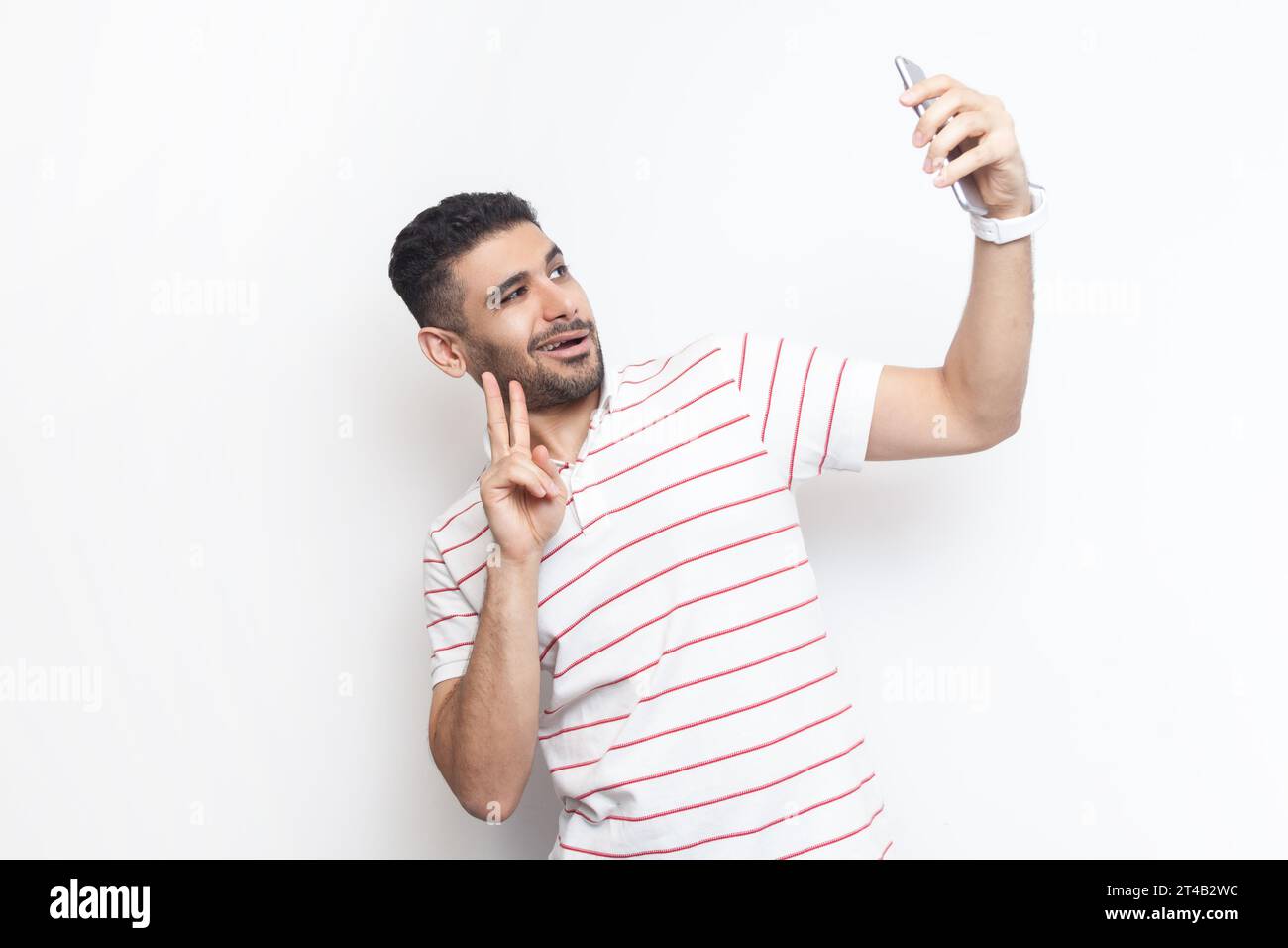 Porträt eines fröhlichen, gutaussehenden, verspielten bärtigen Mannes, der ein gestreiftes T-Shirt trägt und Selfie macht, mit V-Zeichen für seine Anhänger. Innenstudio isoliert auf grauem Hintergrund. Stockfoto