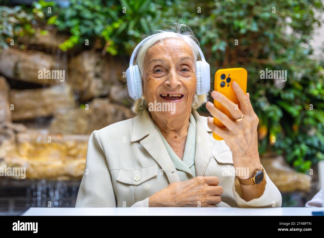 Glückliche ältere Frau mit Kopfhörern und Mobilgeräten, um Musik in einer Geriatrie zu hören Stockfoto