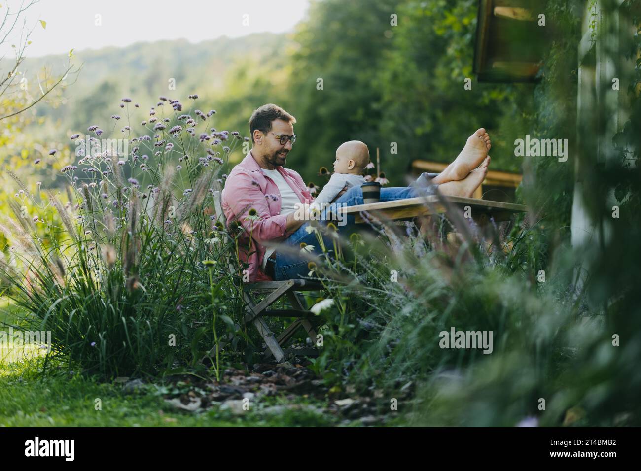 Mann sitzt im Garten mit Füßen auf dem Tisch und spielt mit seinem kleinen Sohn. Vater hat Bindungsmoment mit seinem kleinen Kind. Stockfoto