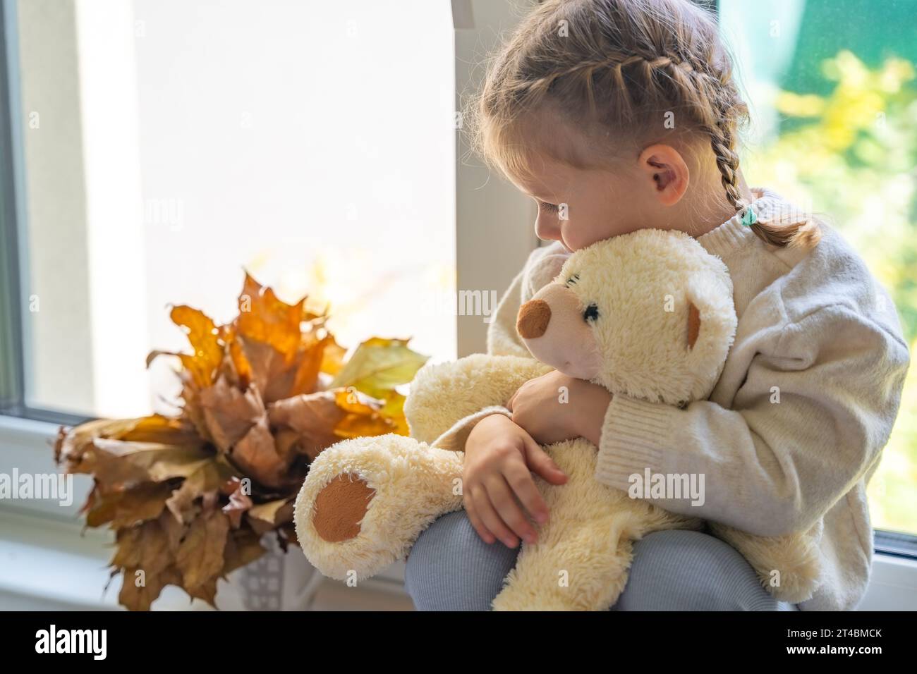 Porträt eines süßen kleinen Mädchens im warmen Pullover, das im Herbst mit einem kleinen Teddybären am Fenster sitzt Stockfoto