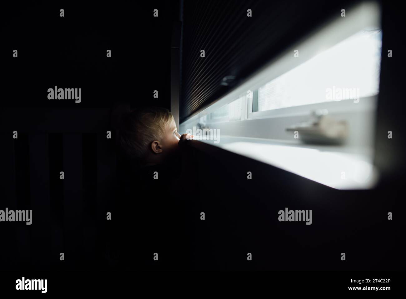 Der kleine blonde Junge ragt durch ein helles Fenster aus dem dunklen Raum Stockfoto