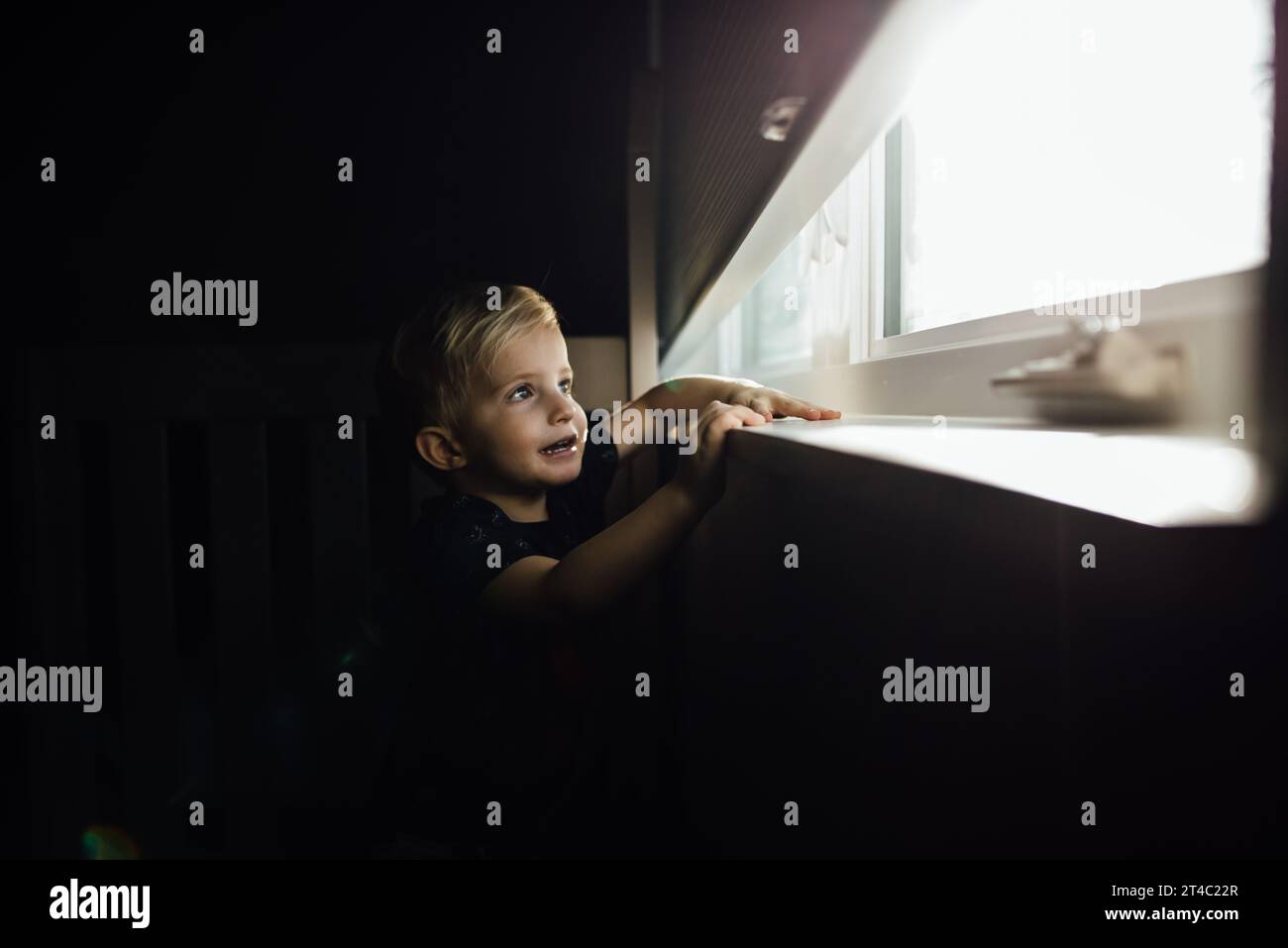 Der kleine blonde Junge schaut durch hellen Wind aus dem dunklen Raum Stockfoto