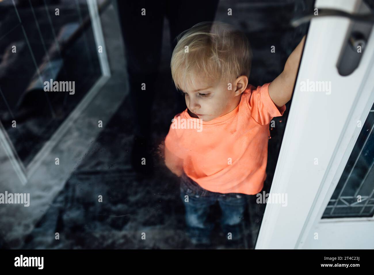 Kleiner Junge in hellem Hemd schaut sehnsüchtig aus der verschmierten Tür nach t Stockfoto