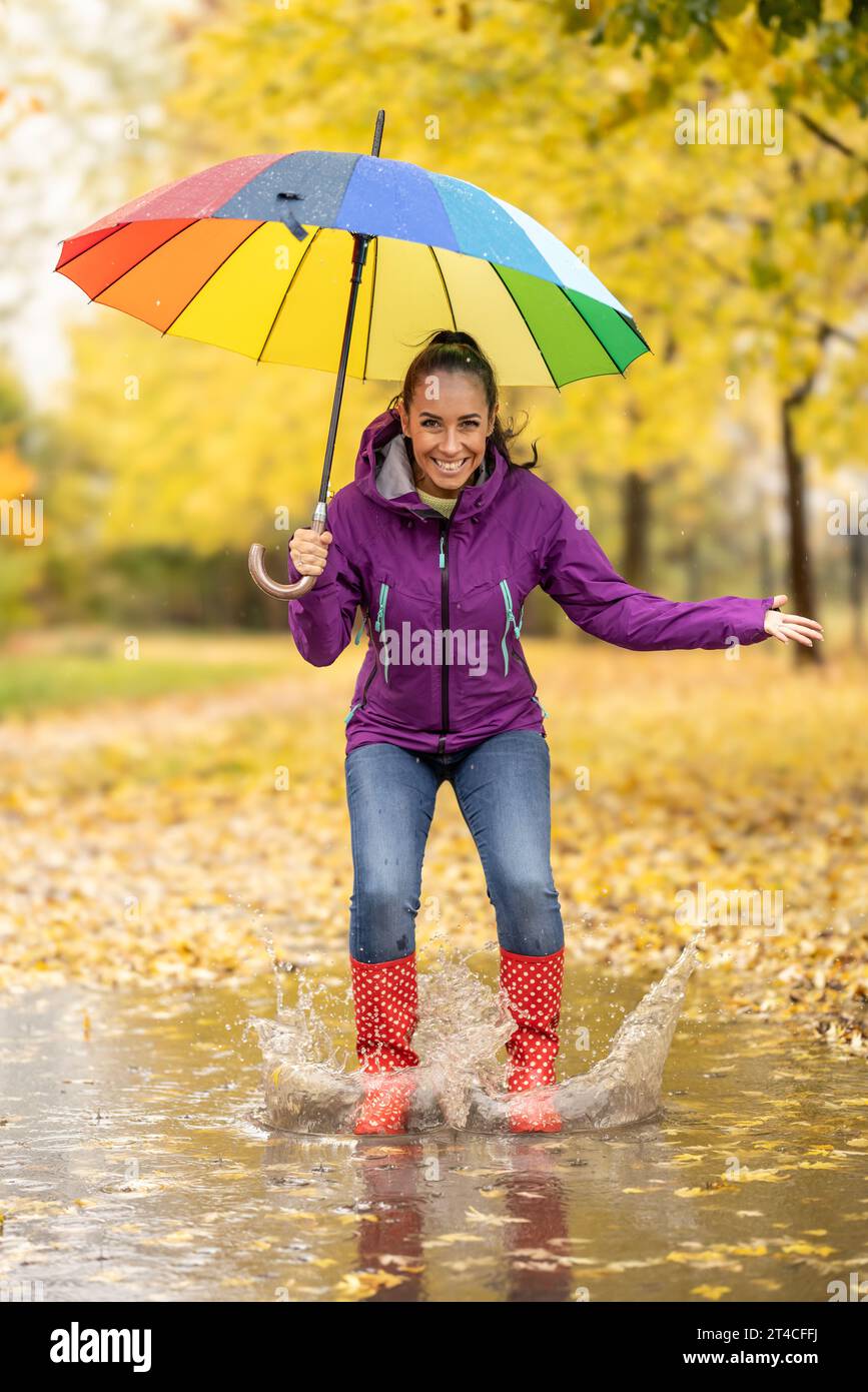 Glückliche junge Frau, die im regnerischen Herbst in Pfützen in Stiefeln springt, lacht und einen Regenschirm über sich hält. Stockfoto