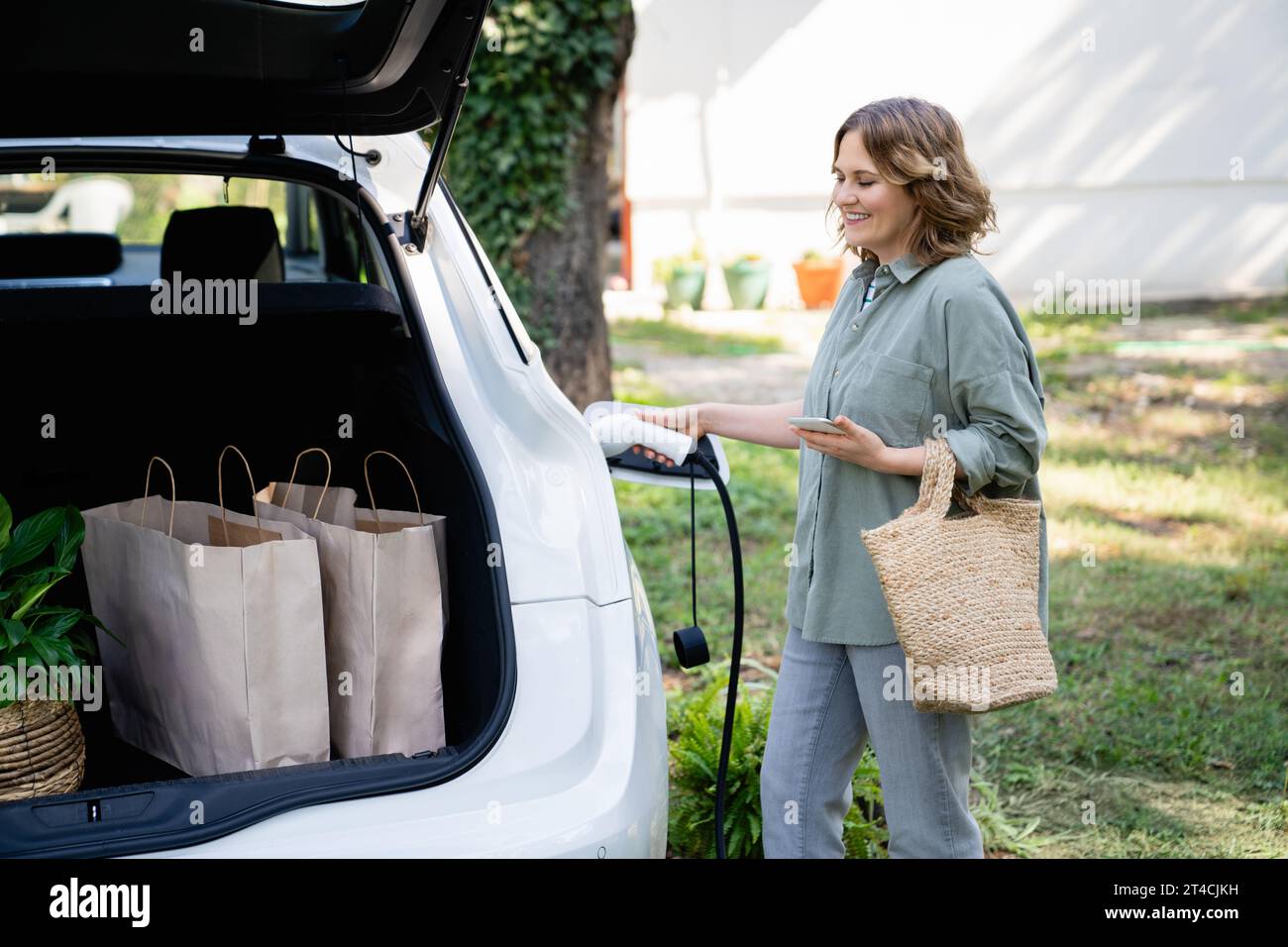 Frau mit Einkaufstasche neben einem aufladenden Elektroauto im Hof eines Landhauses. Stockfoto