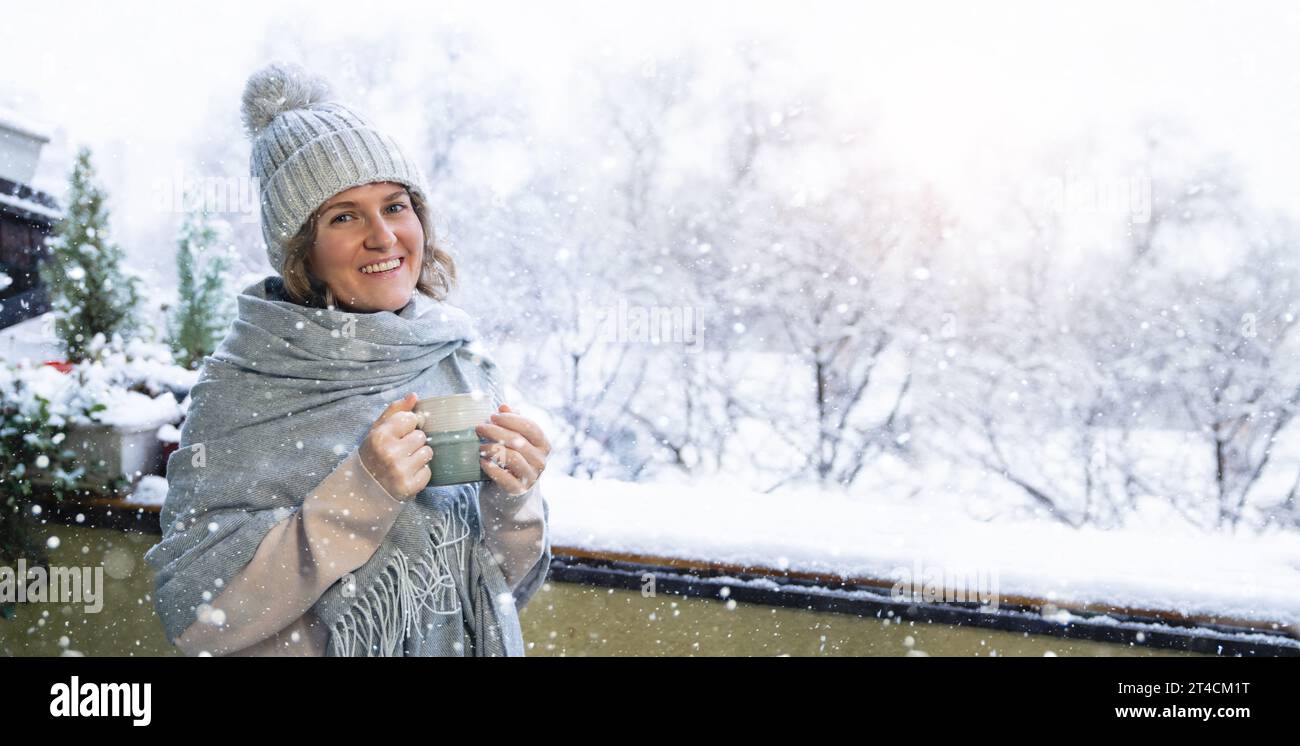 Eine Frau in einem warmen Hut und einer Decke hält einen Becher mit einem heißen Getränk in den Händen. Winterlandschaft mit Schneefall im Hintergrund. Stockfoto