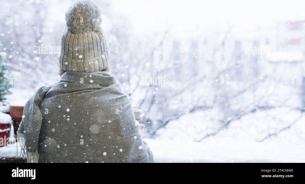 Eine Frau mit warmem Hut und Decke auf der Terrasse. Winterliche Stadtlandschaft mit Schneefall im Hintergrund. Stockfoto