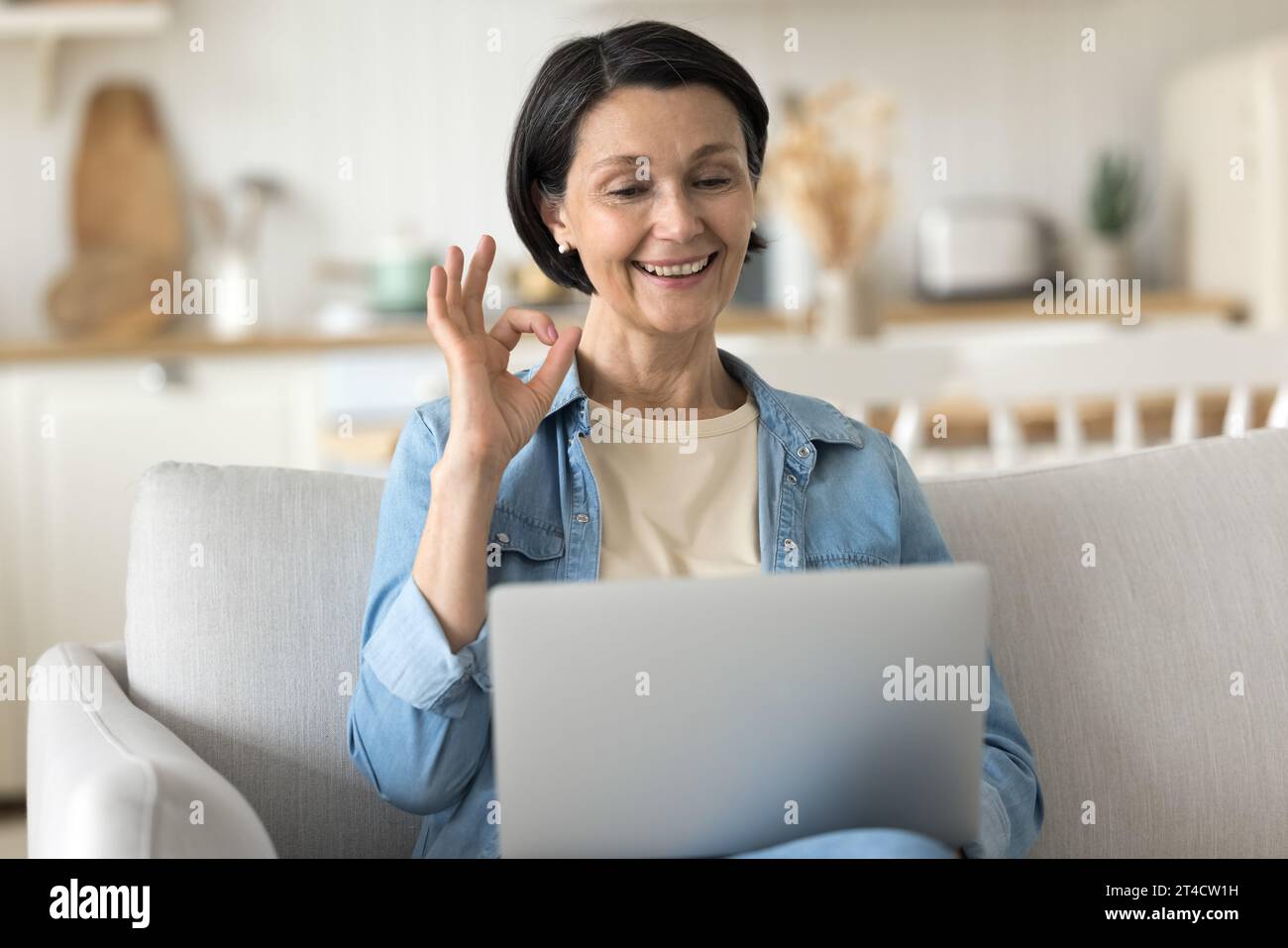 Reife Frau sitzt auf dem Sofa mit Laptop und zeigt eine gute Geste Stockfoto