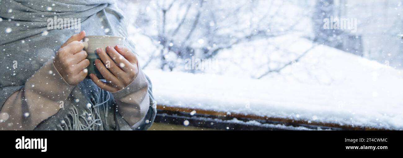 Eine Frau in einem warmen Hut und einer Decke hält einen Becher mit einem heißen Getränk in den Händen. Winterlandschaft mit Schneefall im Hintergrund. Stockfoto