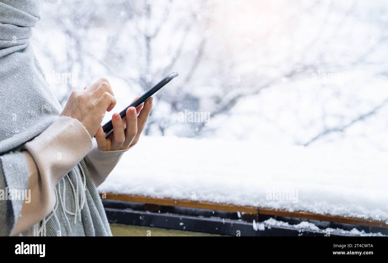 Eine Frau in einem warmen Hut und einer Decke hält das Smartphone in den Händen. Winterlandschaft mit Schneefall im Hintergrund. Stockfoto