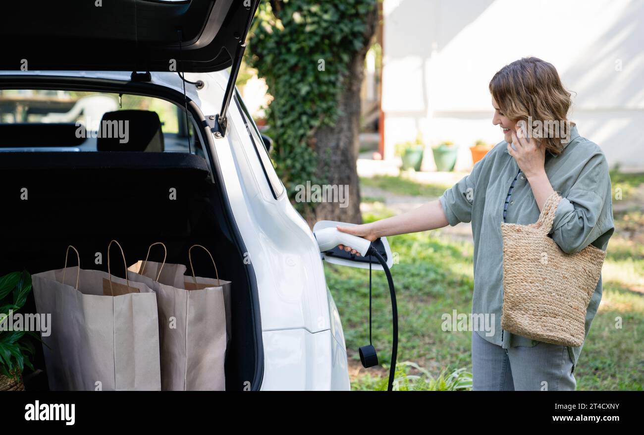 Frau mit Einkaufstasche neben einem aufladenden Elektroauto im Hof eines Landhauses. Stockfoto