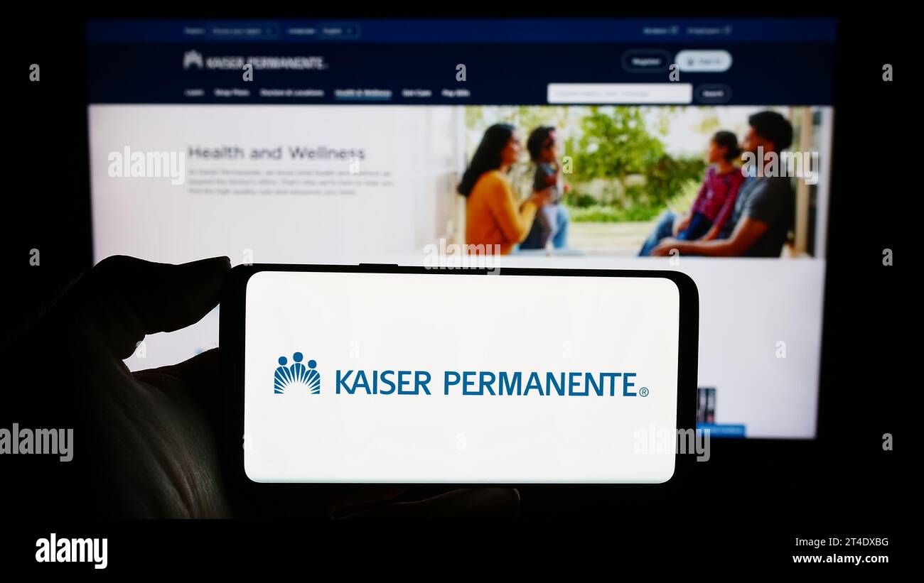 Person mit Mobiltelefon mit Logo des amerikanischen Managed Care-Konsortiums Kaiser Permanente vor der Webseite. Schwerpunkt auf dem Display des Telefons. Stockfoto