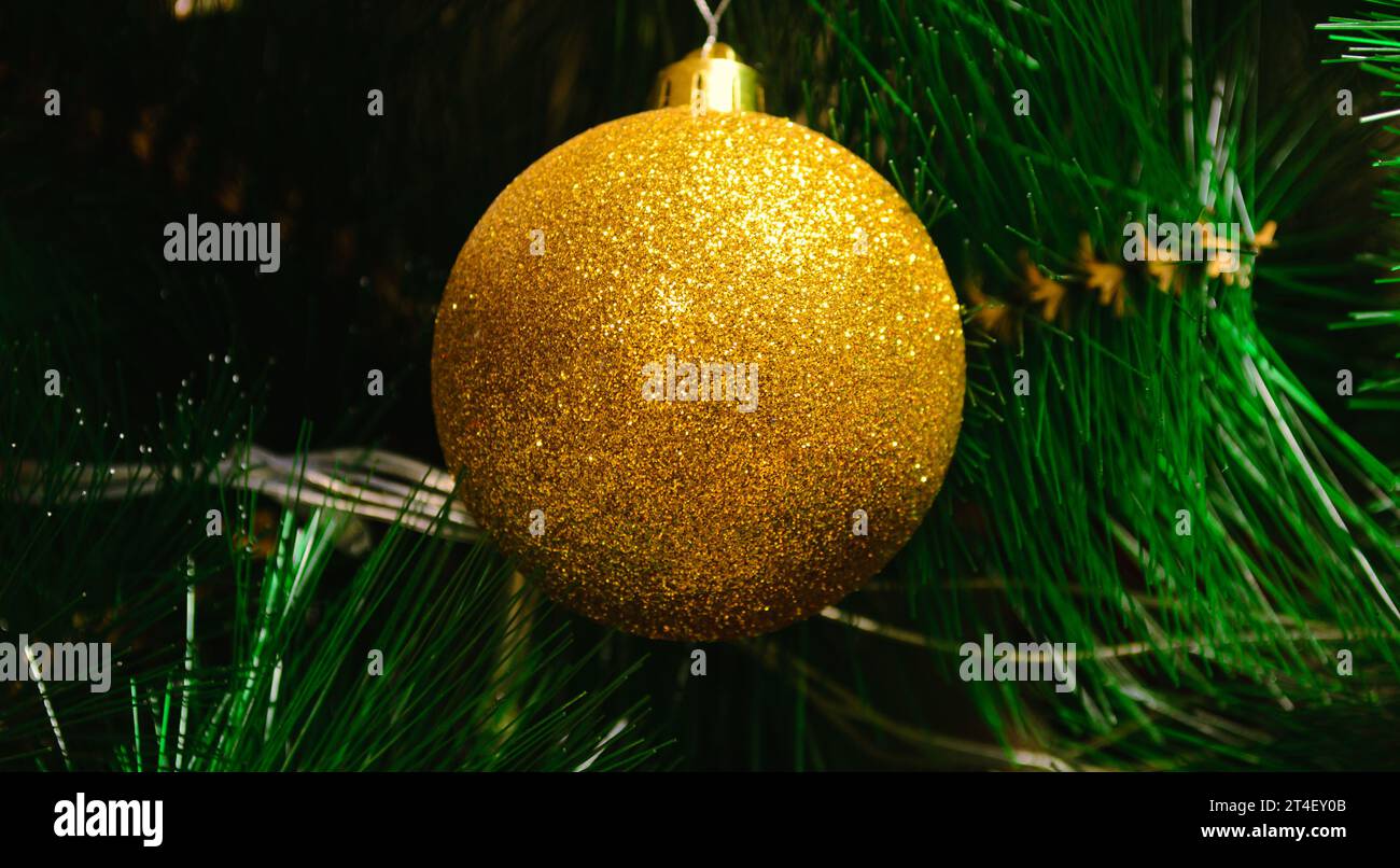 Ein Spielzeug in Form einer goldenen Kugel hängt vor dem Hintergrund grüner Zweige eines künstlichen Weihnachtsbaums Stockfoto