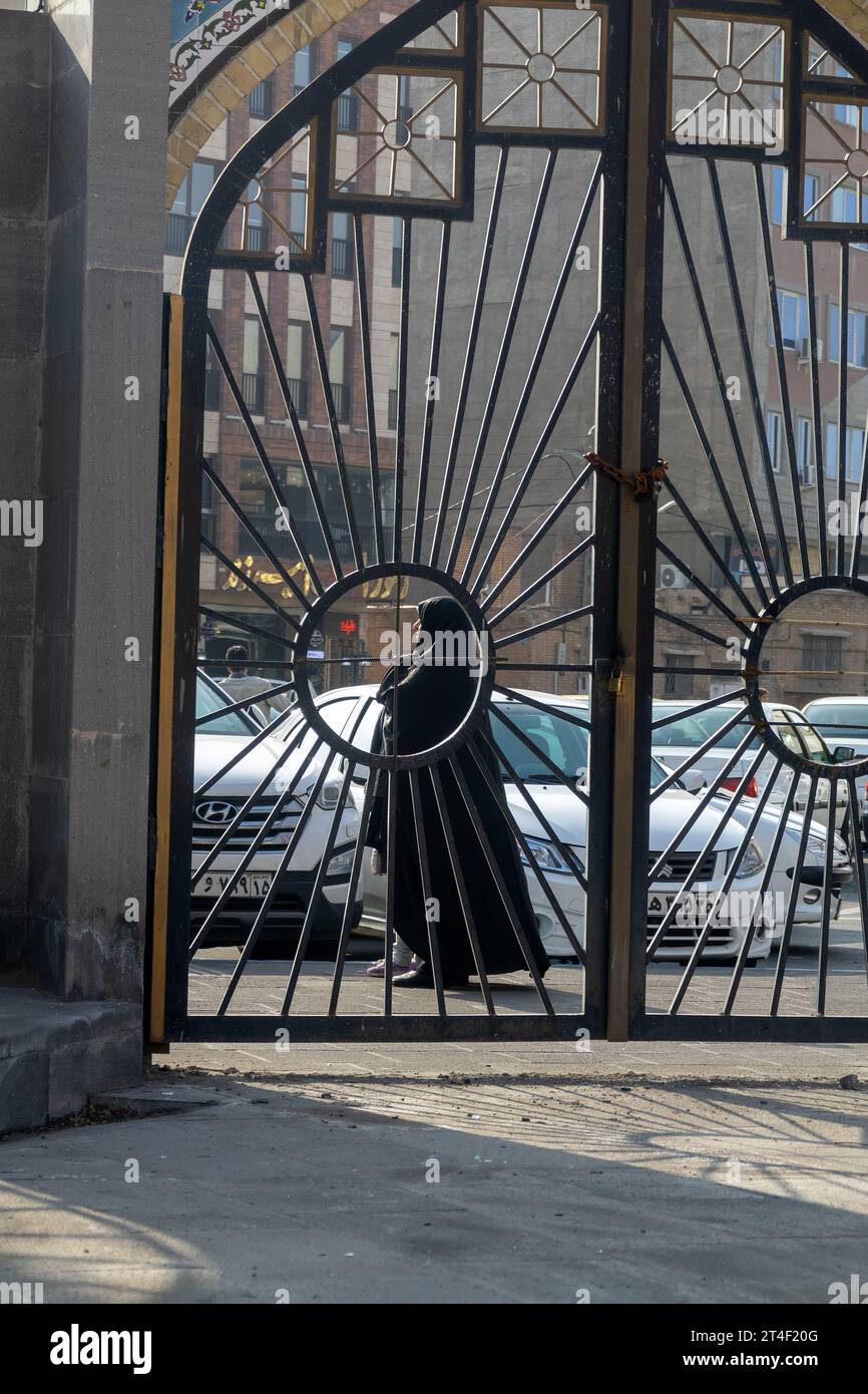 Tabriz, Iran, 14. Dezember 2022: Eine alte iranische Frau in einem schwarzen Schleier hinter einem gemusterten Tor (in einem Gitter aus schwarzen Toren) auf der Straße Stockfoto