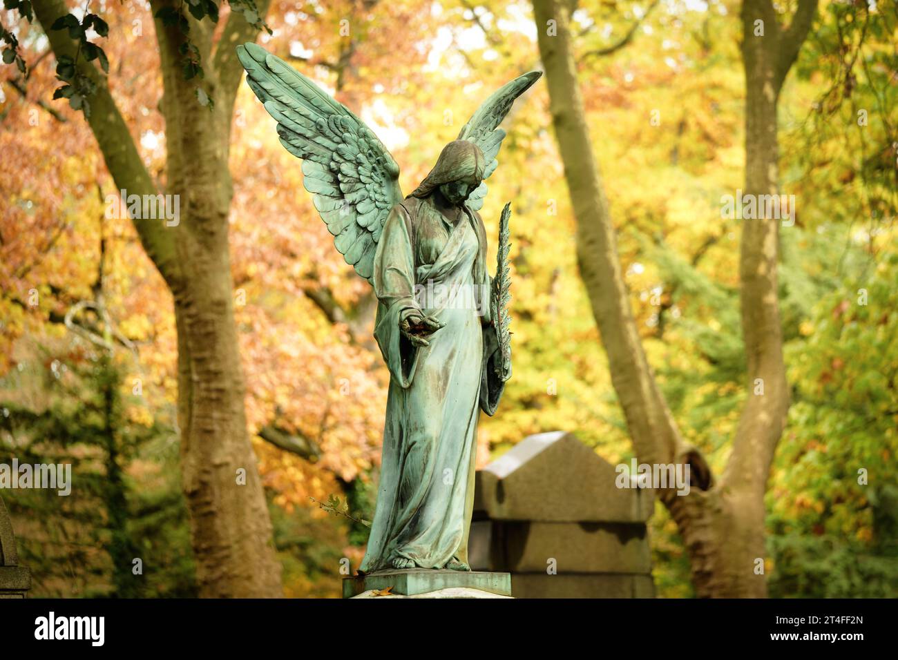 Eine antike Engelsfigur mit Palmenzweig steht auf einem Grab eines Friedhofs vor einem herbstlichen bunten Laubwald in einem verschwommenen Hintergrund Stockfoto