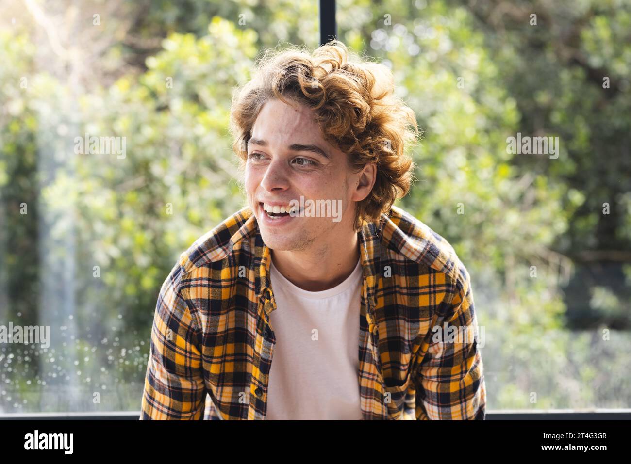 Glücklicher kaukasischer Mann mit lockigen Haaren, der zu Hause am Fenster sitzt Stockfoto