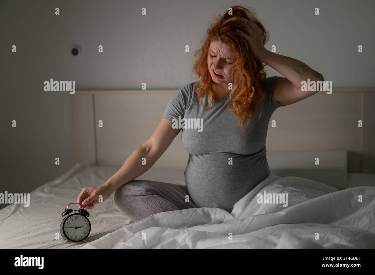 Eine schwangere Frau sitzt im Bett und schaut auf den Wecker. Schlaflosigkeit. Stockfoto